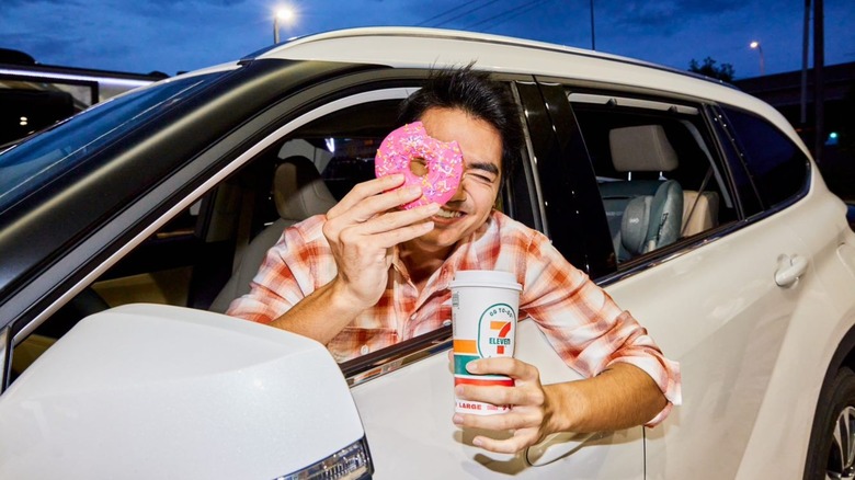 man leaning out of driver's side of car holding pink donut and 7-Eleven coffee