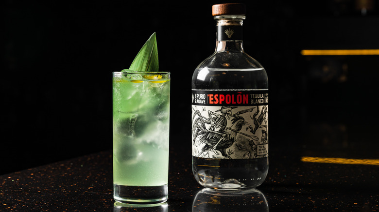 A bottle of Espolòn Blanco next to a glass of mojito on reflective countertop.