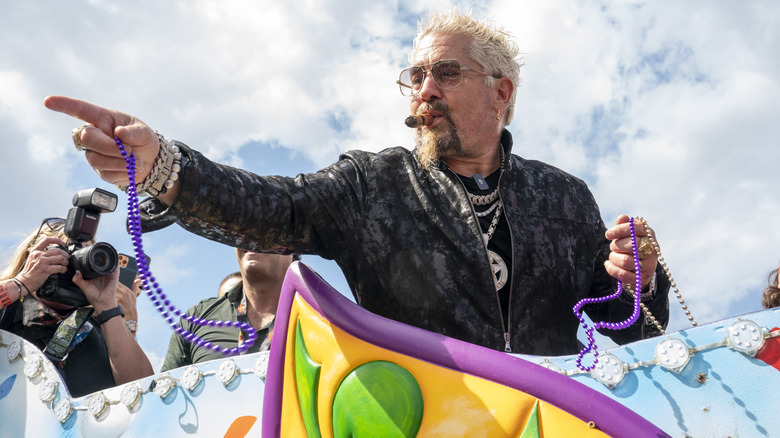 Guy Fieri smoking a cigar on a Mardi Gras float, holding beads and pointing