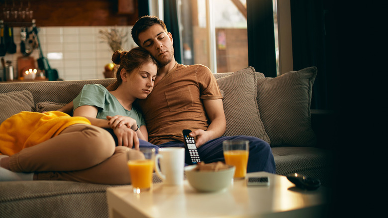 A young couple asleep on the couch while the man holds a TV remote