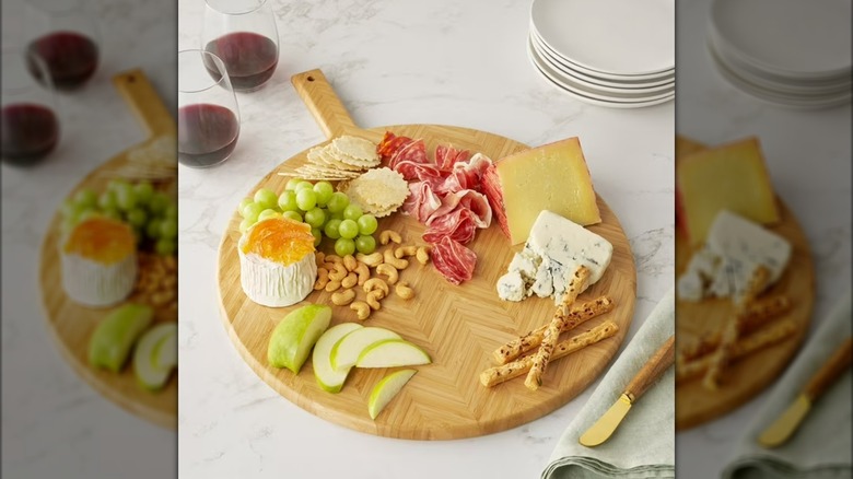 Round bamboo board with fruit, nuts, and cheese on top, with glasses of red wine
