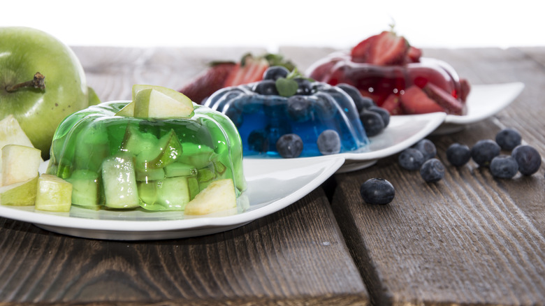 Three different Jell-O desserts on white plates with blueberries, strawberries and green apple on the side