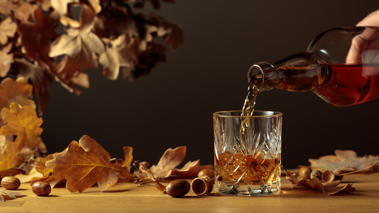 Pouring whiskey from brown bottle into rocks glass with dried oak leaves background