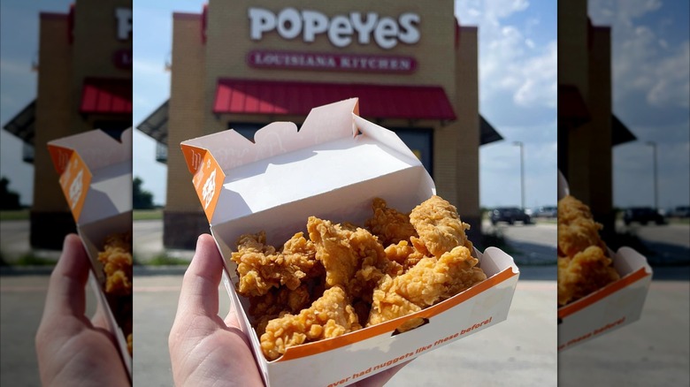 Hand holding up a box of Popeyes chicken nuggets outside of a Popeyes restaurant