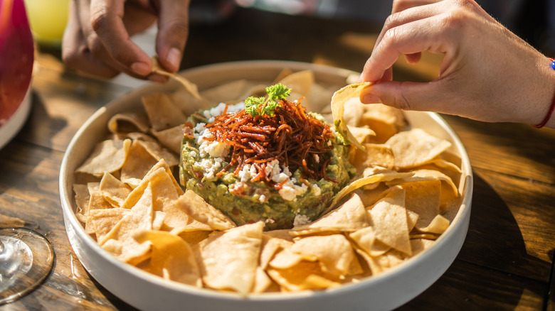Chips and gauc in a white bowl with two hands reaching for a chip
