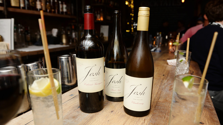 Three bottles of Josh Cellars wine on a wooden bar surrounded by water glasses, lemon, lime, and straws
