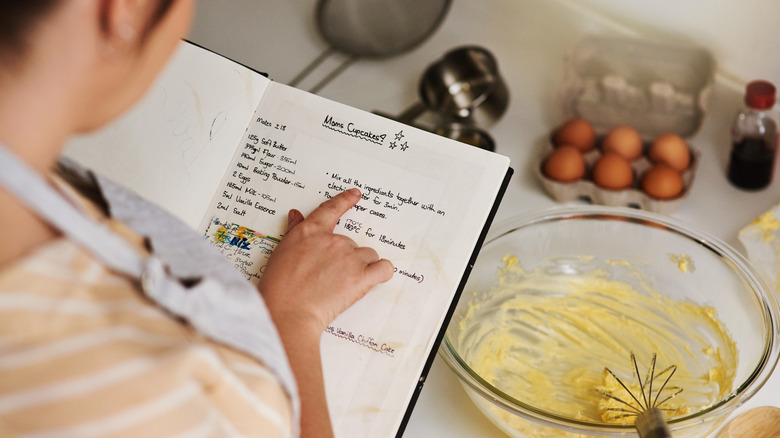 A person reading a handwritten recipe in a notebook with a bowl of cupcake batter, a whisk, measuring cups, and eggs