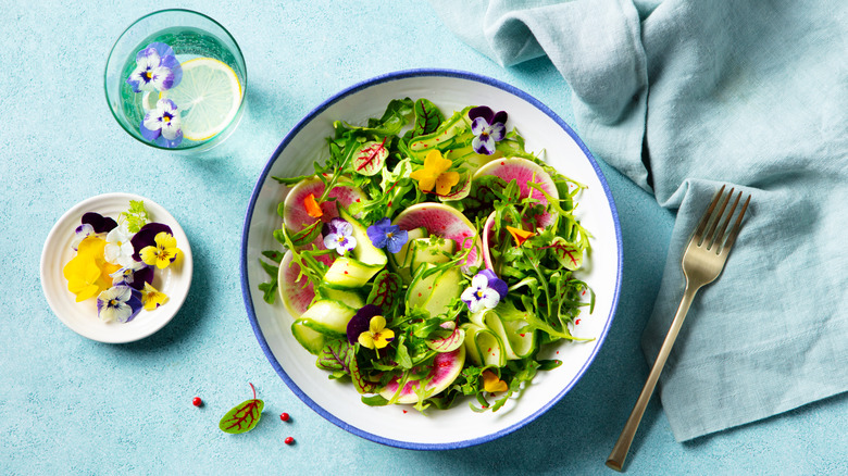 An overhead view of a green salad with sliced watermelon radish, cucumber ribbons, leafy greens, and colorful edible flowers with a fork, napkin, and a glass of lemon water with edible flowers