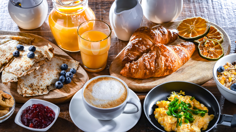 A breakfast spread with croissants, crepes topped with blueberries, scrambled eggs with green onions, yogurt and granola, coffee, and glasses of orange juice