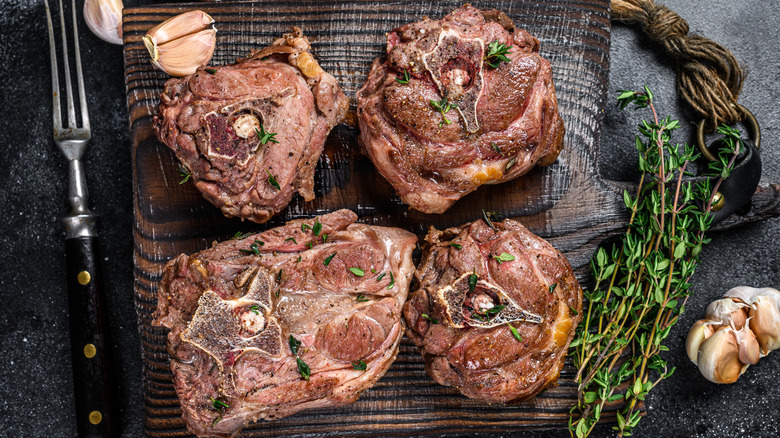 Grilled lamb neck bones on a wooden cutting board with garlic cloves and herbs