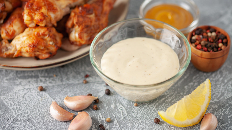 white barbecue sauce in glass bowl next to a plate of grilled chicken drumsticks and seasonings like lemon, black pepper, and garlic