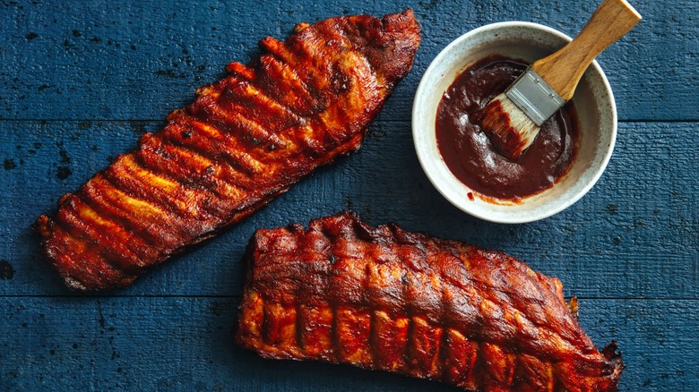 two racks of grilled ribs on table next to bowl of barbecue sauce with brush
