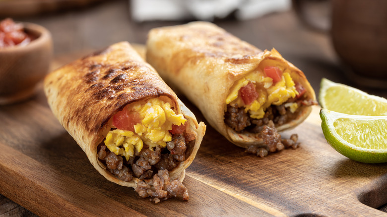 Breakfast burrito filled with eggs, salsa, and sausage sliced in half and placed on a wooden cutting board with lime wedges to garnish