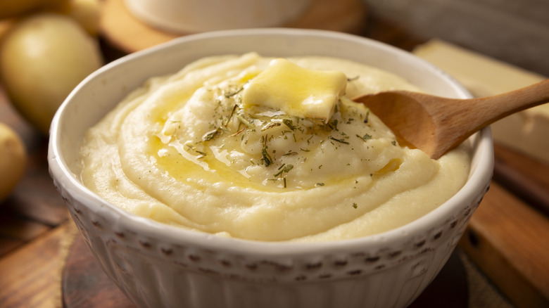 Homemade mashed potatoes topped with a pat of butter and fresh rosemary in a white bowl with a wooden spoon in it