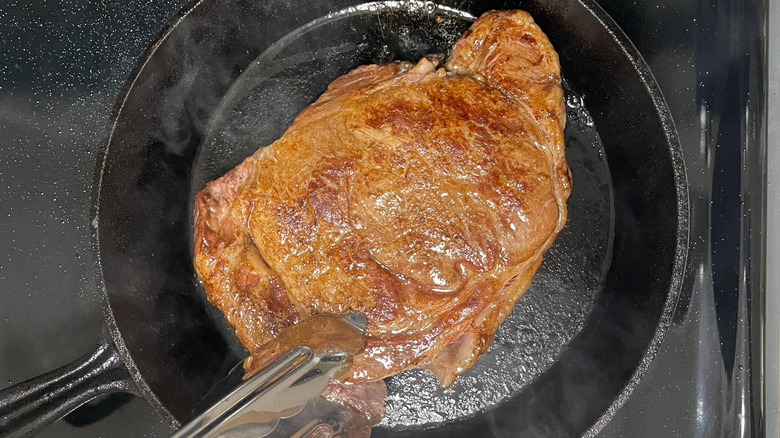 Tongs placing ribeye steak in cast iron skillet