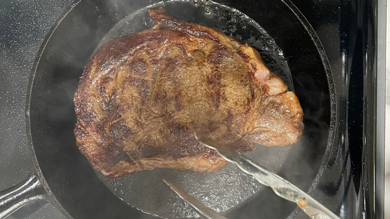 Tongs placing ribeye steak in cast iron skillet