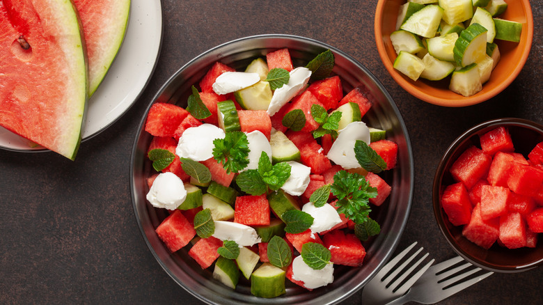 Diced watermelon, cheese, cut cucumber, and leafy garnishes next to slices of watermelon and utensils.