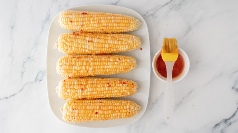 Sauced corn on the cob on plate next to bowl of chili sauce with pastry brush