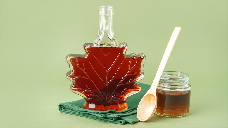 Maple syrup inside maple leaf-shaped bottle and small jar with wooden spoon, against green backdrop.
