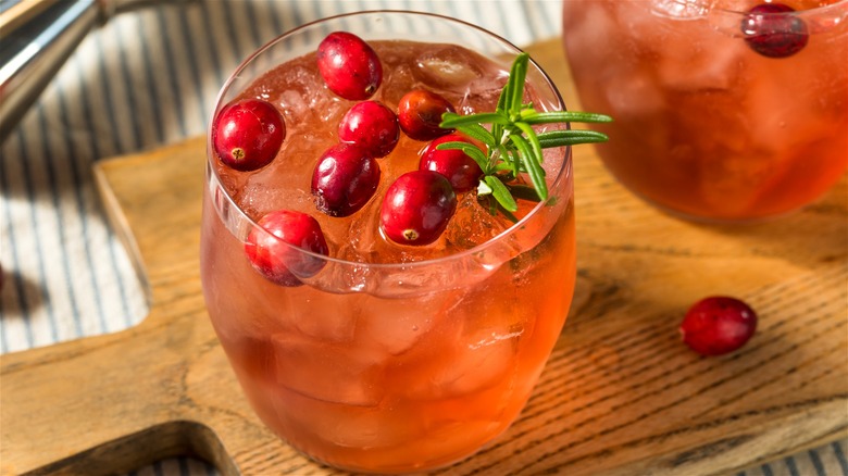 Vodka cranberry cocktail garnished with fresh berries and rosemary sprig, served on a wooden board.