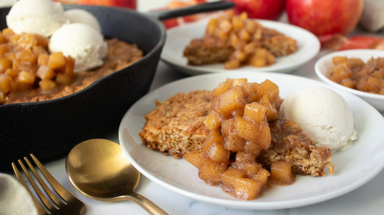 cookie topped with apples and ice cream in a black frying pan and several white dishes