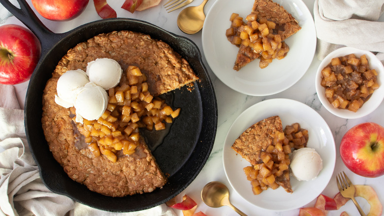 cookie topped with apples and ice cream in a black frying pan and several white dishes