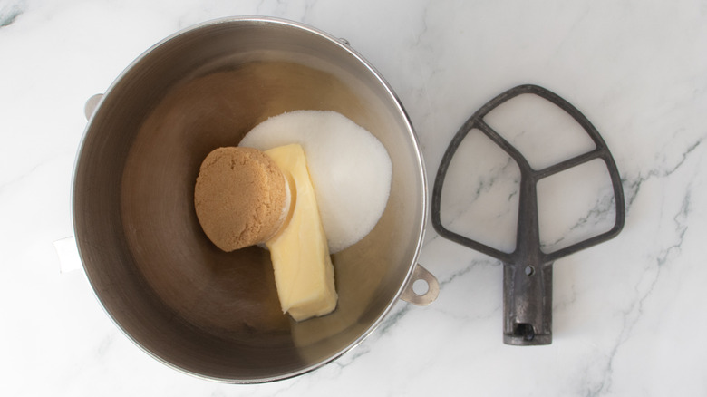 brown sugar, white sugar, and butter in a metal bowl next to the paddle attachment for a stand mixer