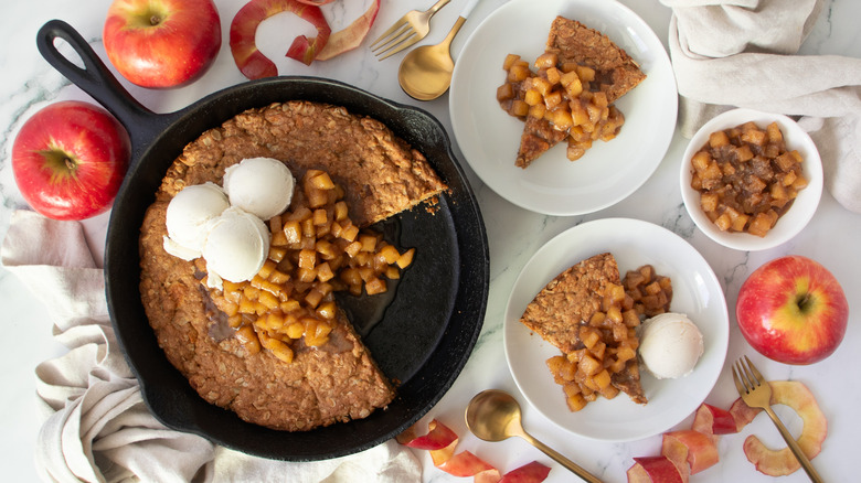 cookie topped with apples and ice cream in a black frying pan and several white dishes
