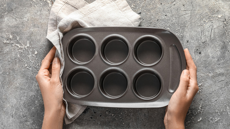 Hands holding a muffin tin with a cloth on a stone surface