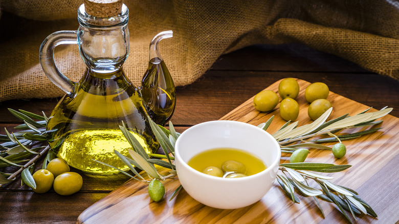 Olive oil in small white bowl on wooden board with olives, glass bottles, and a burlap sack nearby
