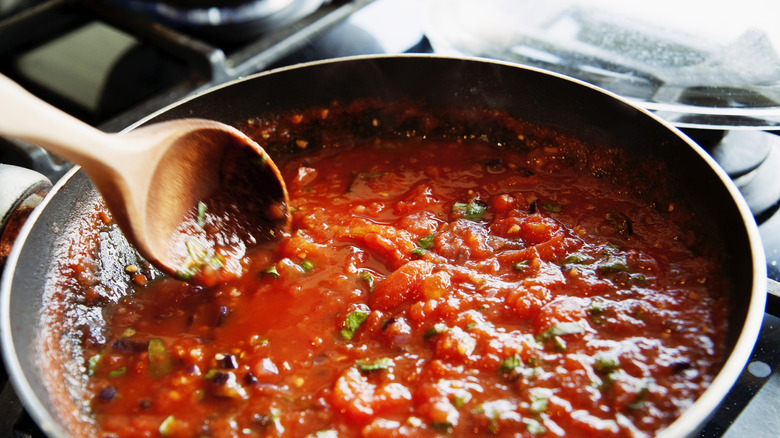 A wooden spoon mixes a mixture of tomatoes and herbs on a pan on the stove.