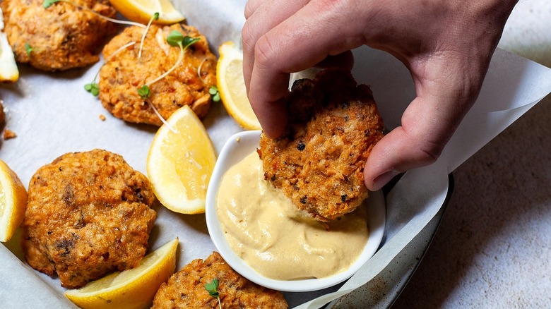 A salmon patty dipping into a sauce with lemon wedges on the side