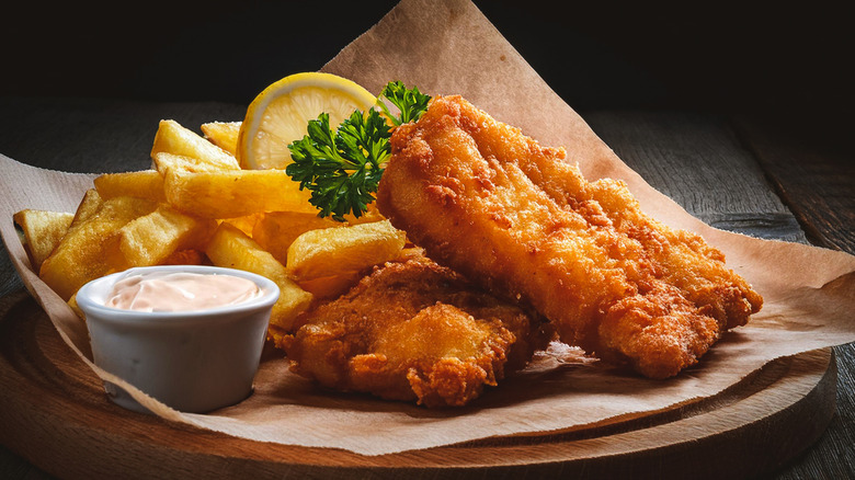 Fried fish, fries, sauce, lemon and parsley on parchment paper