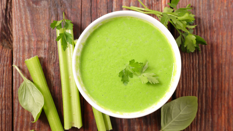 White bowl of green soup with celery on wood surface