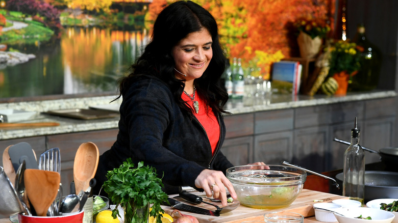 Alex Guarnaschelli wearing a red shirt, black jacket doing a cooking demonstration