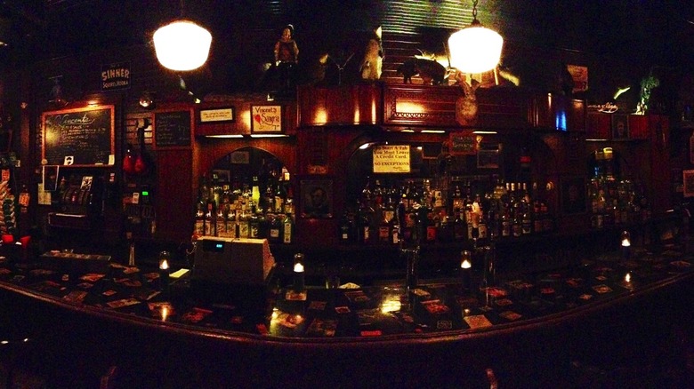 Inside of a dimly lit bar with a cash register, bottles and a counter.