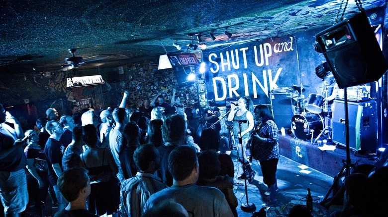 people enjoying live music inside a bar with a sign on the back that reads "shut up and drink""