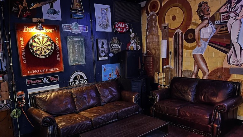 inside of a bar with 2 couches, a dart board and beer logos on one wall, and a painting of a woman in the other.