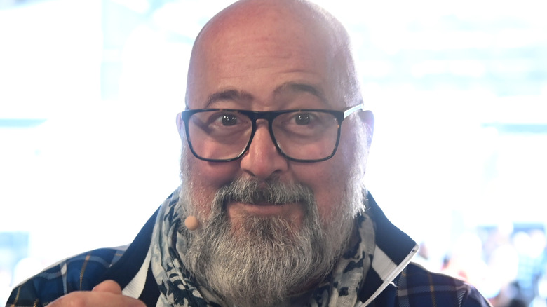 A bearded Andrew Zimmern wears glasses and a microphone