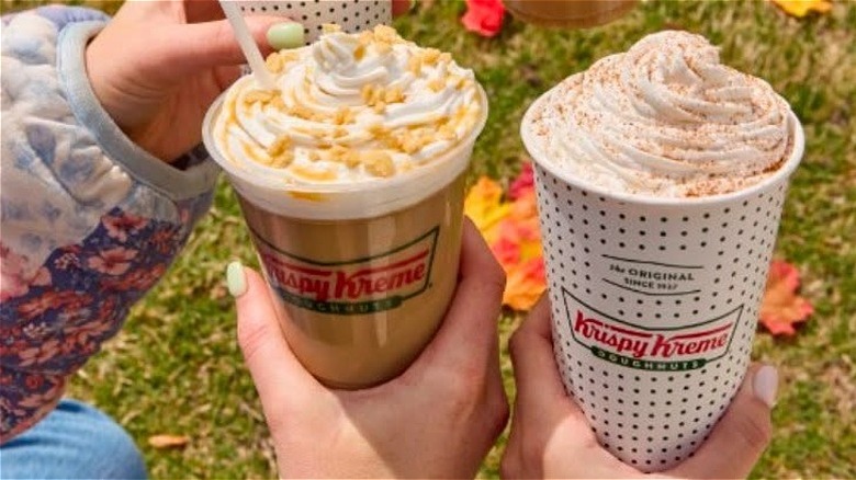 Hands holding two Krispy Kreme coffee beverages on an autumnal leaf-and-grass background