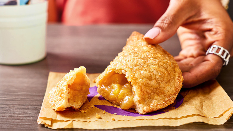A hand holding a Taco Bell caramel apple empanada on a brown paper bag