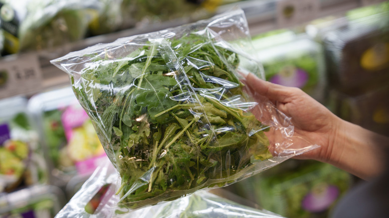 A hand holding a clear plastic bag filled with salad greens inside a grocery store