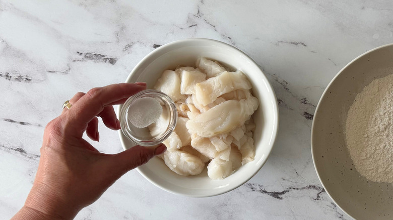 Hand adding salt to cod in bowl