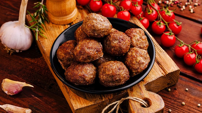 A bowl of well-crusted meatballs served on a wooden plank alongside tomatoes, garlic, pepper, and rosemary.