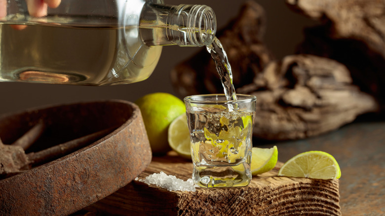 Pouring tequila into a shot glass with limes and a little pile of salt nearby.