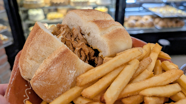 A bifana pork sandwich on a molete roll and french fries with cakes and pastries in the background