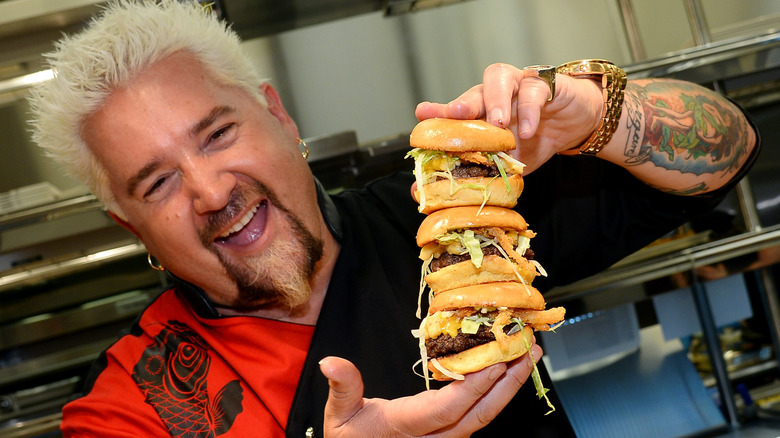 Guy Fieri smiling while holding three burgers squished together in a kitchen