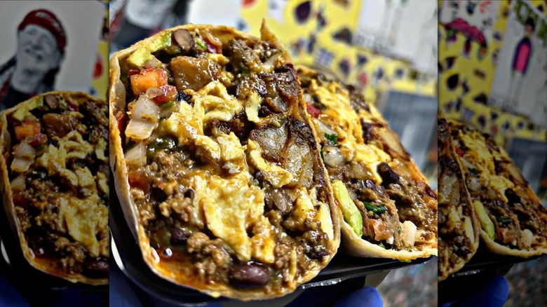 A close-up of a breakfast burrito cut in half packed with eggs, beans, pico de gallo, and avocado with a graffiti-type wall in the background