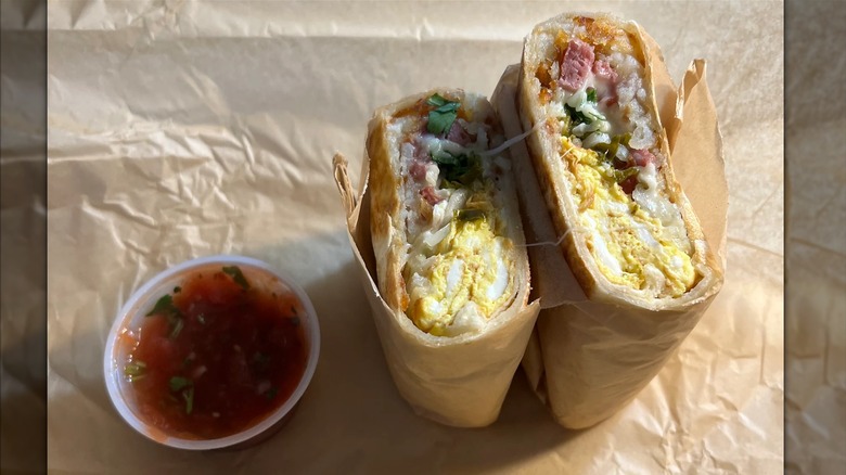 A burrito stuffed with eggs, hashbrowns, cilantro, and reindeer sausage cut in half on a brown paper wrapper with a plastic tub of salsa next to it