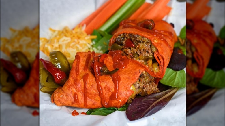 The Kay's 3 Alarm Burrito from Mitchell's Breakfast Burritos, featuring a red tortilla with jalapeños, green chiles, banana peppers, and spicy-hot Italian sausage inside. The burrito is on a plate surrounded by greens, carrot and celery sticks, shredded cheese, and peppers